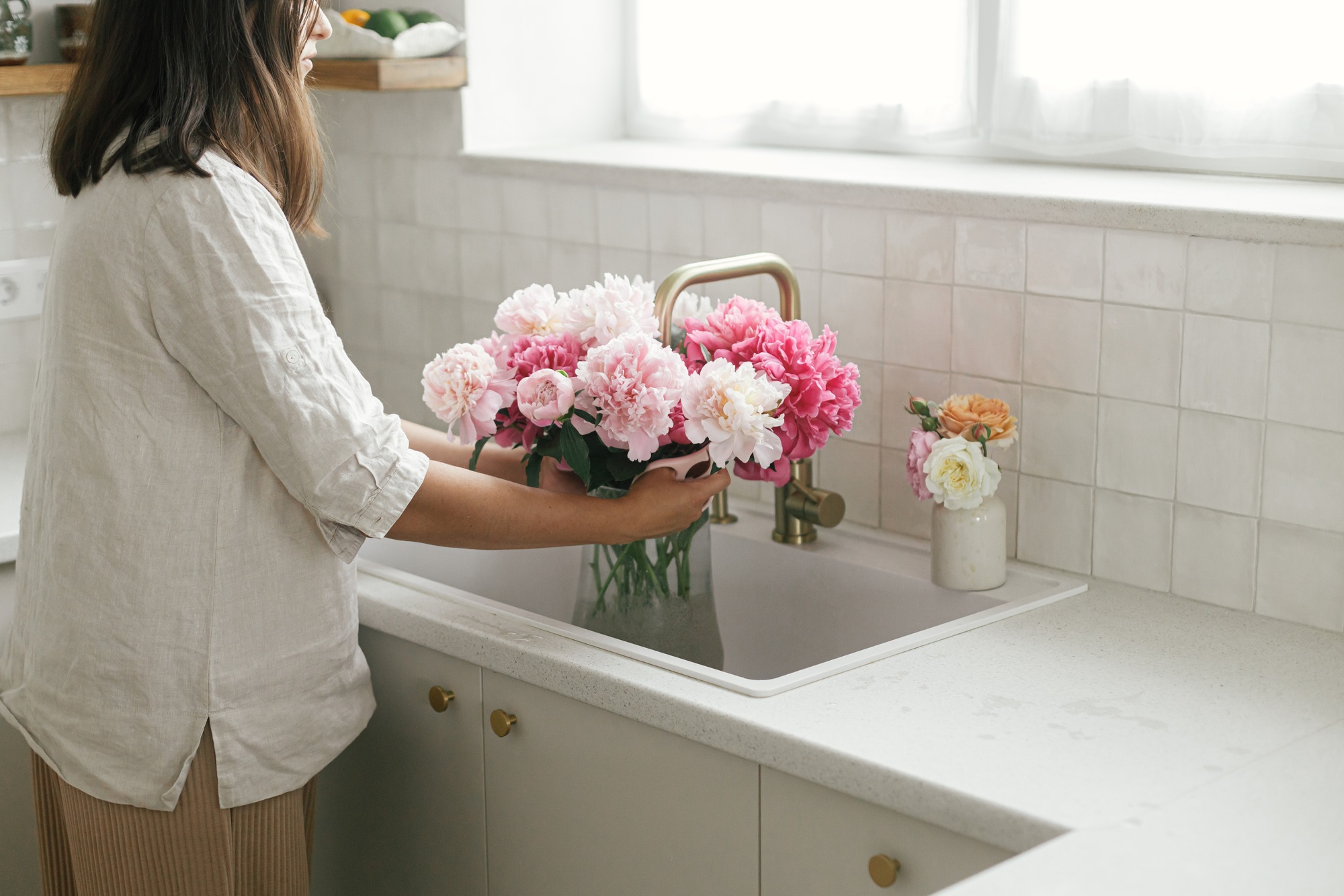 Woman arranging beautiful peonies in vase at sink with brass faucet and granite countertop